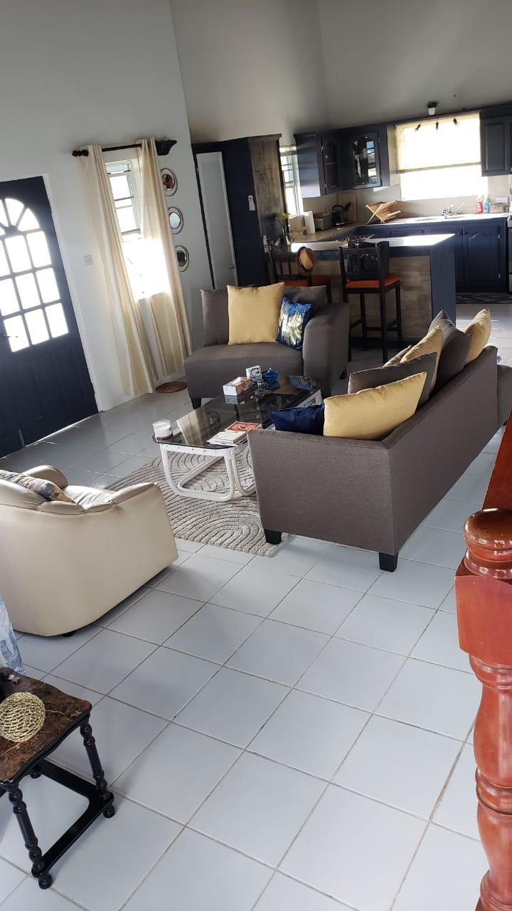 A living area features a neutral-colored sofa set with yellow accent pillows, arranged around a glass coffee table. A patterned area rug lies beneath. The space leads into a kitchen area with dark cabinetry, visible through an open layout illuminated by natural light from windows.