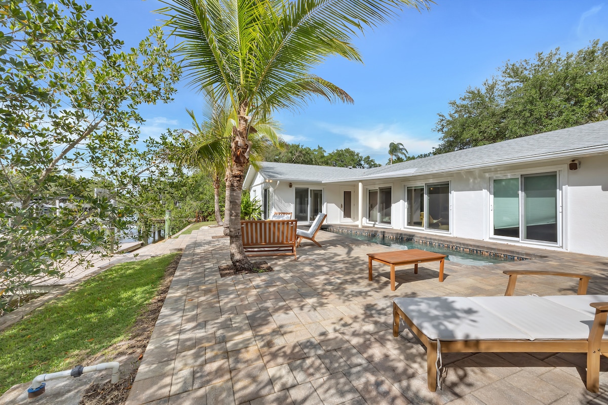 A spacious outdoor area features a swimming pool surrounded by a tiled patio, complemented by lounge chairs and shaded by palm trees. The house is visible in the background with large windows allowing views of the exterior landscape.
