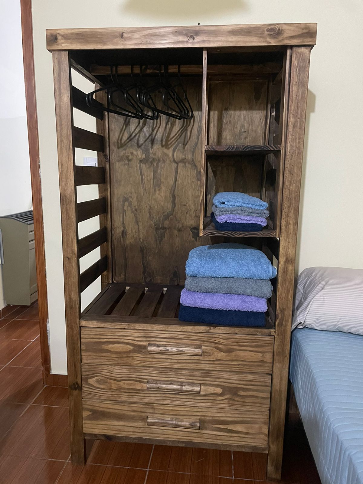 A wooden wardrobe features an open section with hangers and shelves. Neatly stacked towels in various colors rest on a shelf, while two drawers at the bottom offer additional storage. The space beside the wardrobe includes a bed with a light-colored bedspread.