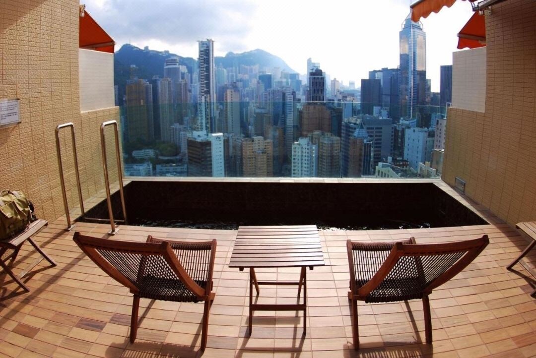 A rooftop terrace features wooden lounge chairs and a small table, overlooking a skyline of tall buildings and mountains in the distance. A swimming pool edge creates a seamless connection to the panoramic view, with bright orange awnings providing shade.