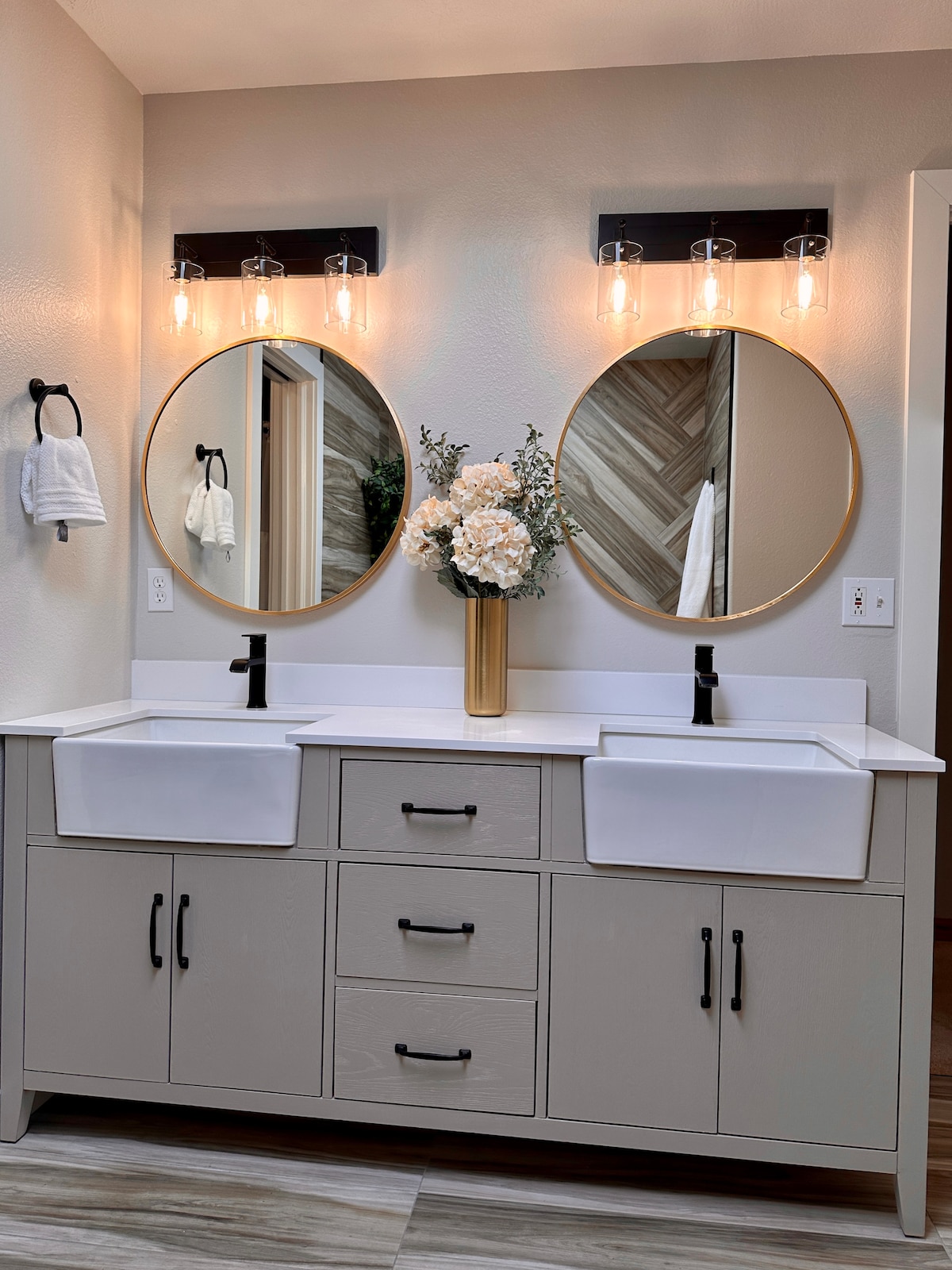 A well-appointed bathroom features dual sinks mounted on a light-colored vanity with multiple drawers. Two round mirrors above the sinks enhance the space, while a tall vase with white flowers adds a touch of elegance. Soft lighting is provided by fixtures on each side.