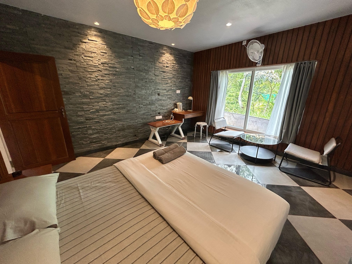 A stylish room features a spacious bed with neutral bedding, complemented by a modern light fixture overhead. A workspace with a wooden desk and comfortable seating is visible near large windows that offer views of the garden. Natural light enhances the room's elegant decor.