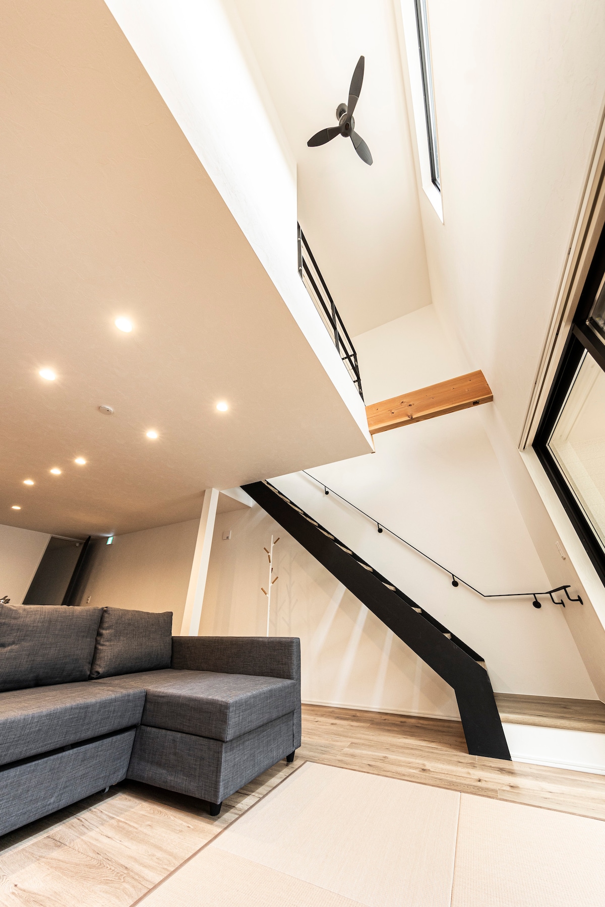 A modern living space features a sectional sofa and a staircase leading to a loft area. Recessed lighting illuminates the high ceiling, while large windows provide natural light. The neutral tones create a calm and open atmosphere.