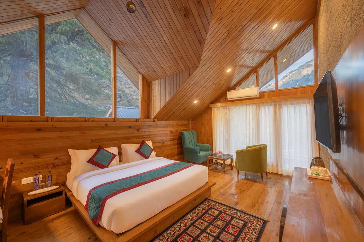A spacious cottage room is highlighted by wooden paneling and an angled ceiling. Two large windows provide natural light and mountain views. A comfortable bed with decorative pillows is positioned alongside a small seating area featuring green chairs. A television and wooden table are visible against the wall.