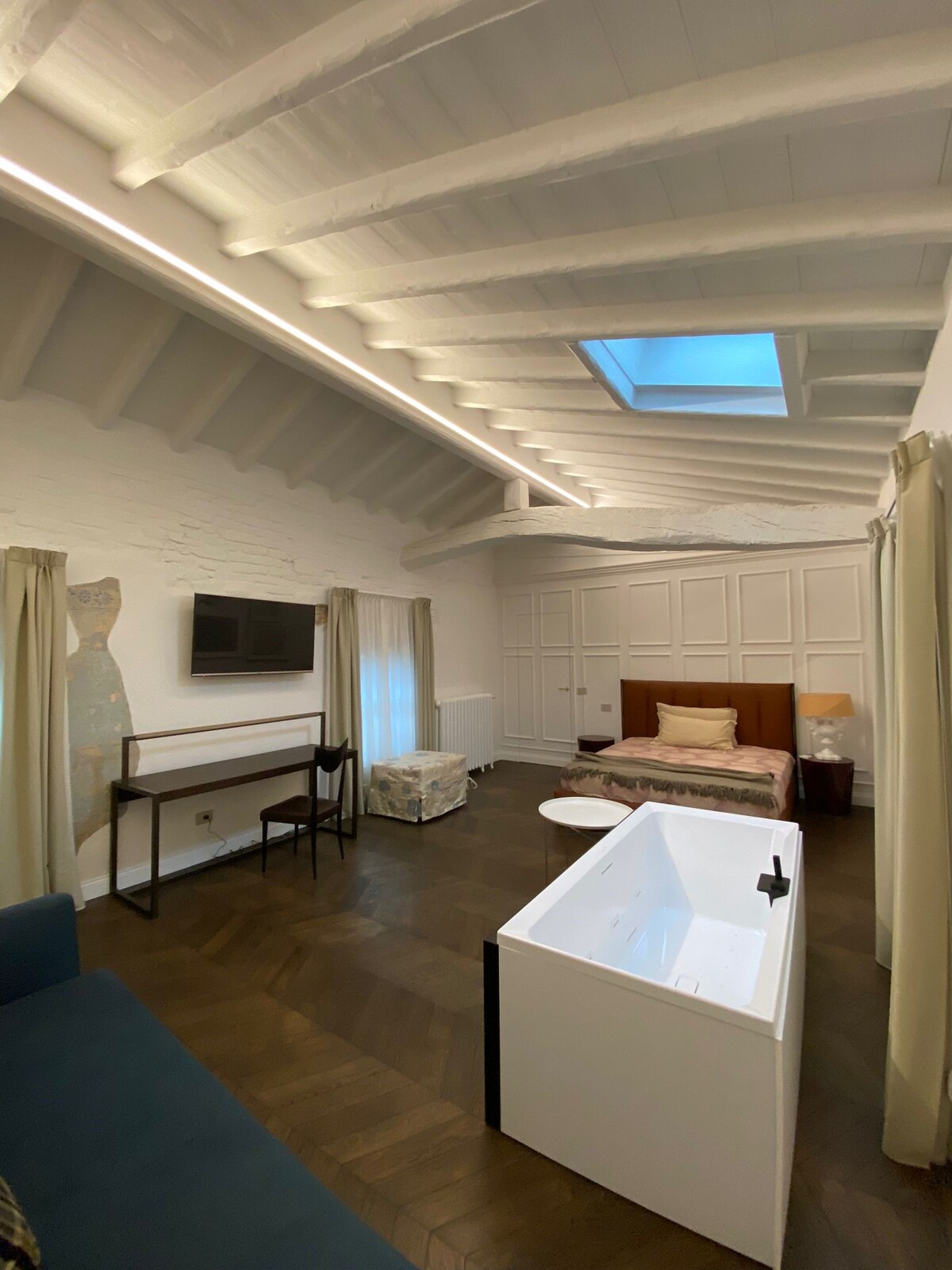 A spacious room is illuminated by natural light from a skylight, highlighting warm wood flooring and high ceiling beams. A cozy sleeping area includes a neatly made bed, while a small seating area features a dark sofa. A minimalist desk and television are also visible.