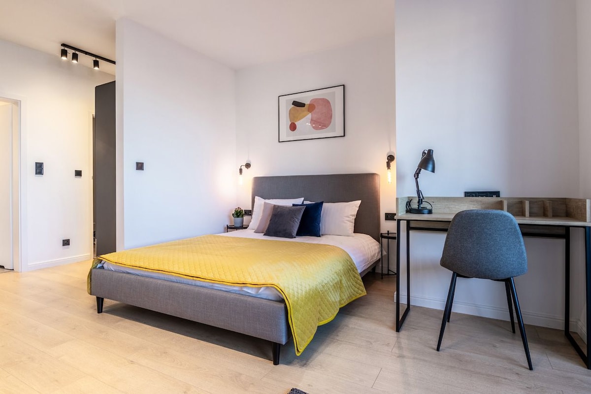 A modern studio bedroom features a spacious bed with a yellow blanket and an assortment of decorative pillows. A small desk and chair are positioned against the wall. Soft lighting from wall sconces creates a warm ambiance.