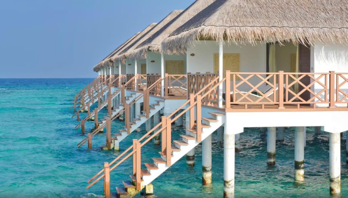 The water villas are positioned on stilts over the turquoise water, featuring thatched roofs and wooden balconies. Steps lead down from each villa, providing access to the sea. The serene blue waters stretch toward the horizon beneath a clear sky.