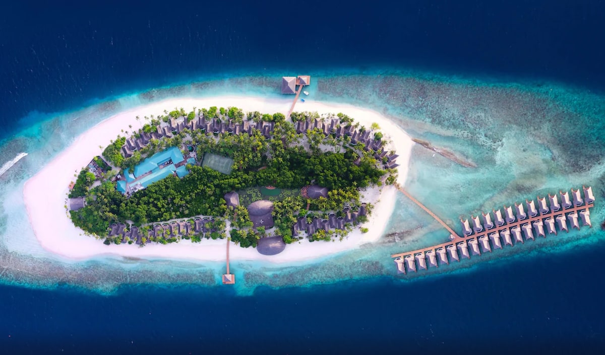 An aerial view showcases a tropical island surrounded by vibrant blue waters. The resort features lush greenery, sandy beaches, and a series of overwater villas extending into the ocean. A pool and various structures are visible among the palm trees, emphasizing the resort's resort-like layout.