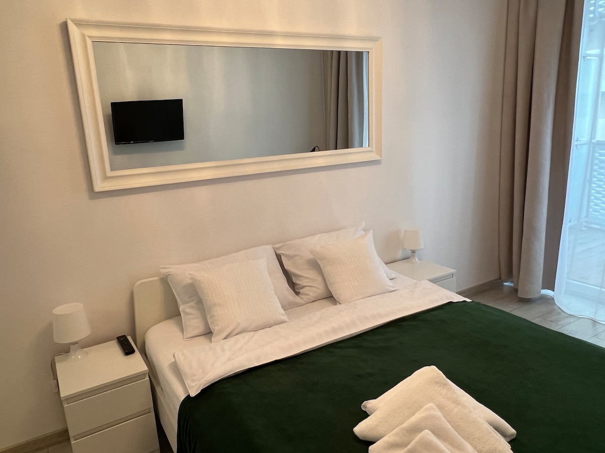A comfortable sleeping area is presented, featuring a spacious bed with white linens and a green throw blanket. Nightstands with lamps are located on either side. A mirror above the bed reflects the light from a nearby window, enhancing the overall brightness of the room.