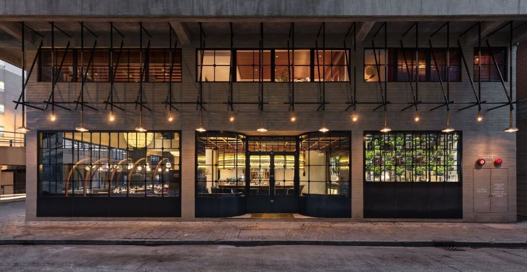 The exterior of a modern building is displayed, featuring large windows with black frames and illuminated interior spaces. The entrance is flanked by outdoor seating areas, and warm lighting from pendant fixtures creates an inviting atmosphere in the evening.