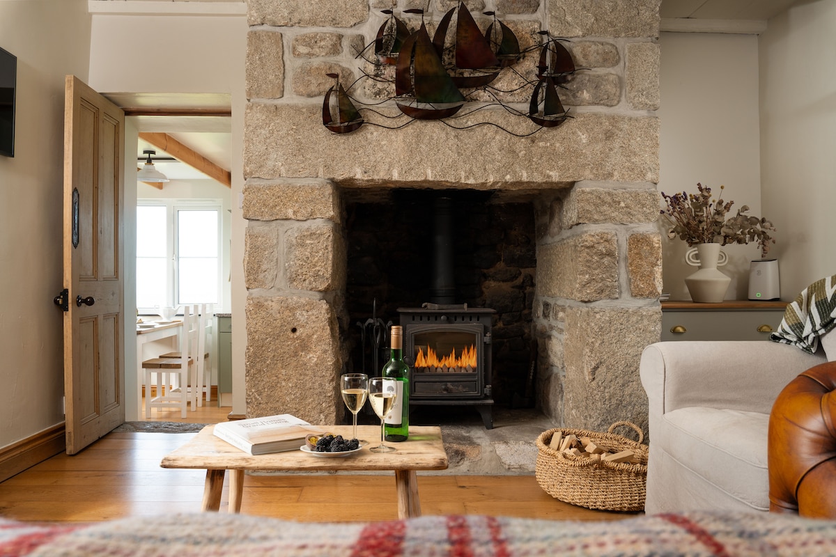 A cozy living area features a stone fireplace with a wood burner, topped with decorative metal sailboat art. A wooden coffee table sits in front, adorned with a bottle of wine and glasses, alongside a basket. Soft textures and natural light enhance the inviting atmosphere.