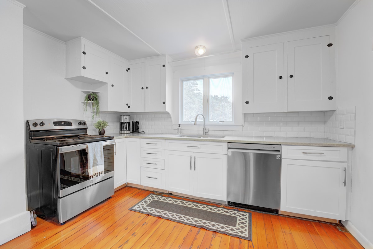 The kitchen showcases white cabinetry with black hardware, complemented by a stainless steel stove and dishwasher. Natural light floods in through the window above the sink, illuminating the countertops. A patterned runner adds a touch of style to the warm wood flooring.