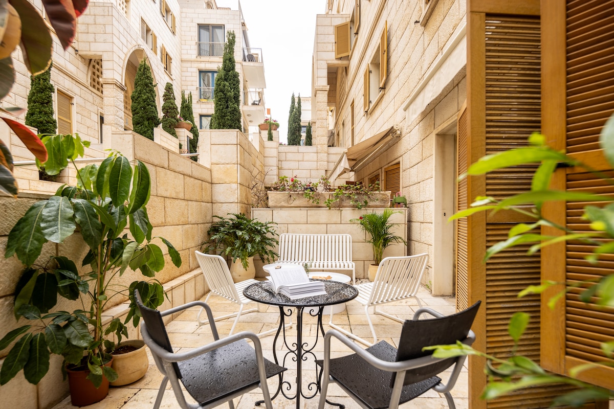 A private outdoor patio is surrounded by low stone walls and greenery. White lounge chairs are arranged around a round table, inviting relaxation. Potted plants add natural beauty, while the space is illuminated by soft, diffused light filtering through nearby buildings.