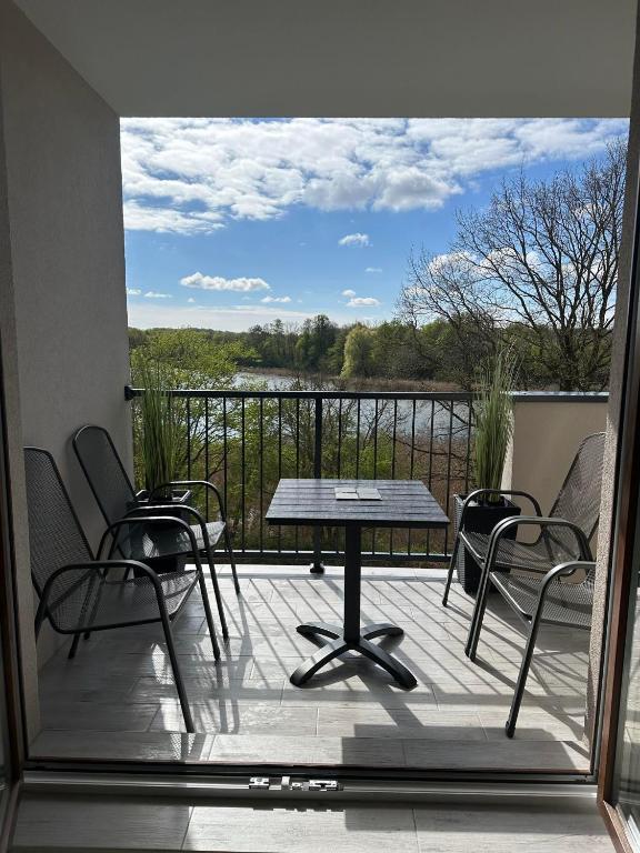 A balcony area features a small table surrounded by four black metal chairs. Green plants decorate the space, with a view of the river and trees in the background under a partly cloudy sky.