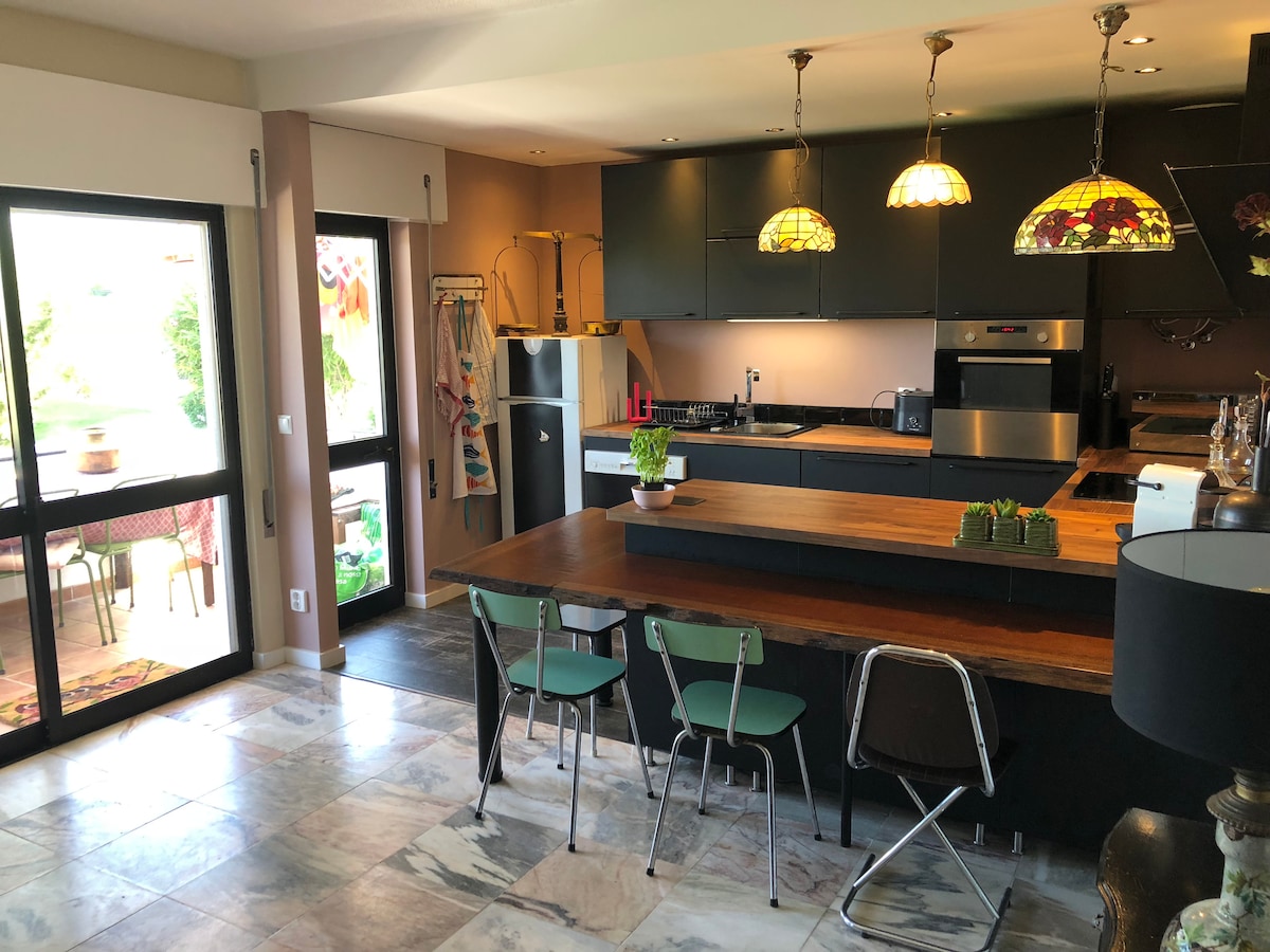 An open-concept kitchen and dining area is presented, featuring modern black cabinetry and a spacious wooden island. Three metal chairs surround the island. Light fixtures with stained glass accents add color. Large glass doors open to a balcony view, enhancing the connection to the outdoors.