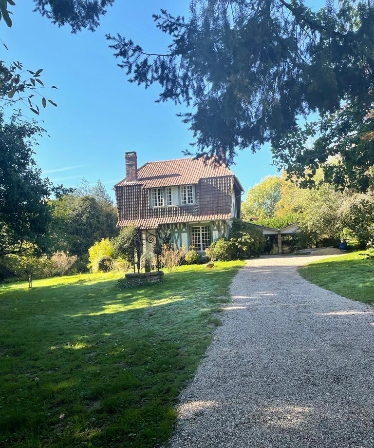 A charming cottage-style house is set among lush greenery, featuring a tiled roof and large windows. A gravel pathway leads to the entrance, surrounded by well-maintained gardens. The sky above is clear with a gentle blue hue, enhancing the serene rural ambiance.