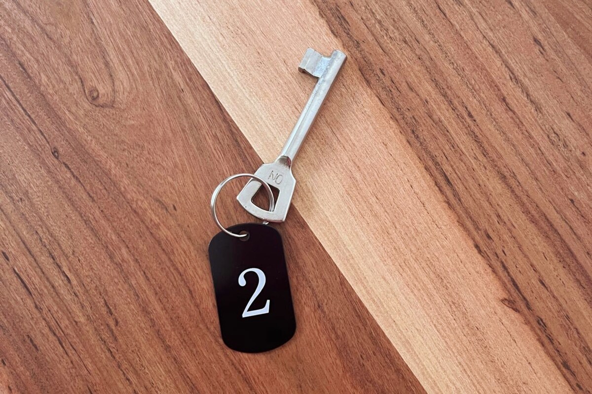 A metallic key rests on a wooden surface, accompanied by a black tag displaying the number '2.' The wood's texture showcases a blend of light and dark tones, highlighting the natural grain.