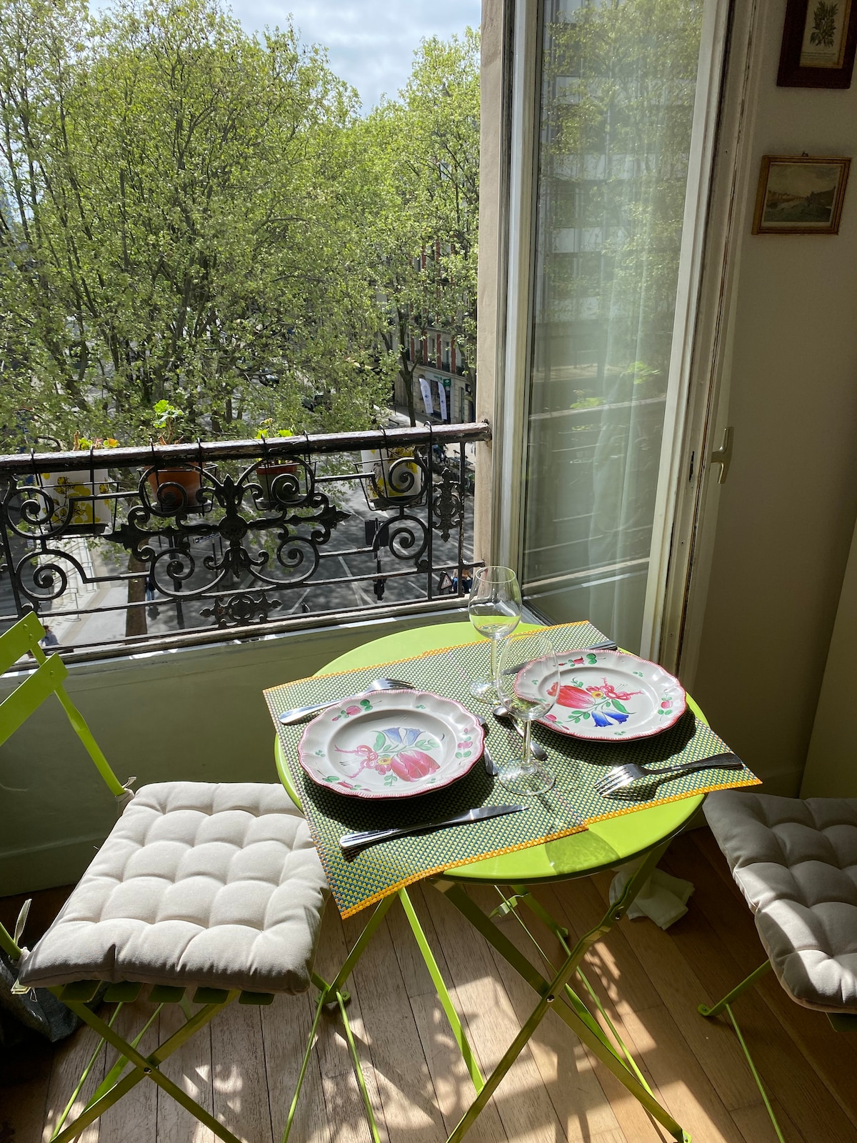 A small dining table is set for two, featuring colorful plates and a glass. The table is positioned near a window with leafy trees visible outside. Two cushioned chairs are placed at the table, creating a cheerful dining space.