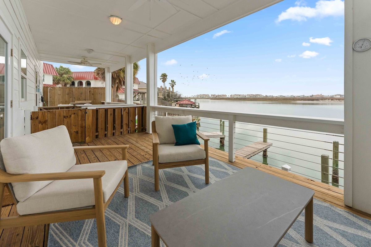 An outdoor patio area features two light-colored chairs paired with a coffee table, offering a view of the waterfront. The wooden railing adds a natural touch, and the surrounding scenery includes palm trees and water. A rug with geometric patterns enhances the inviting space.