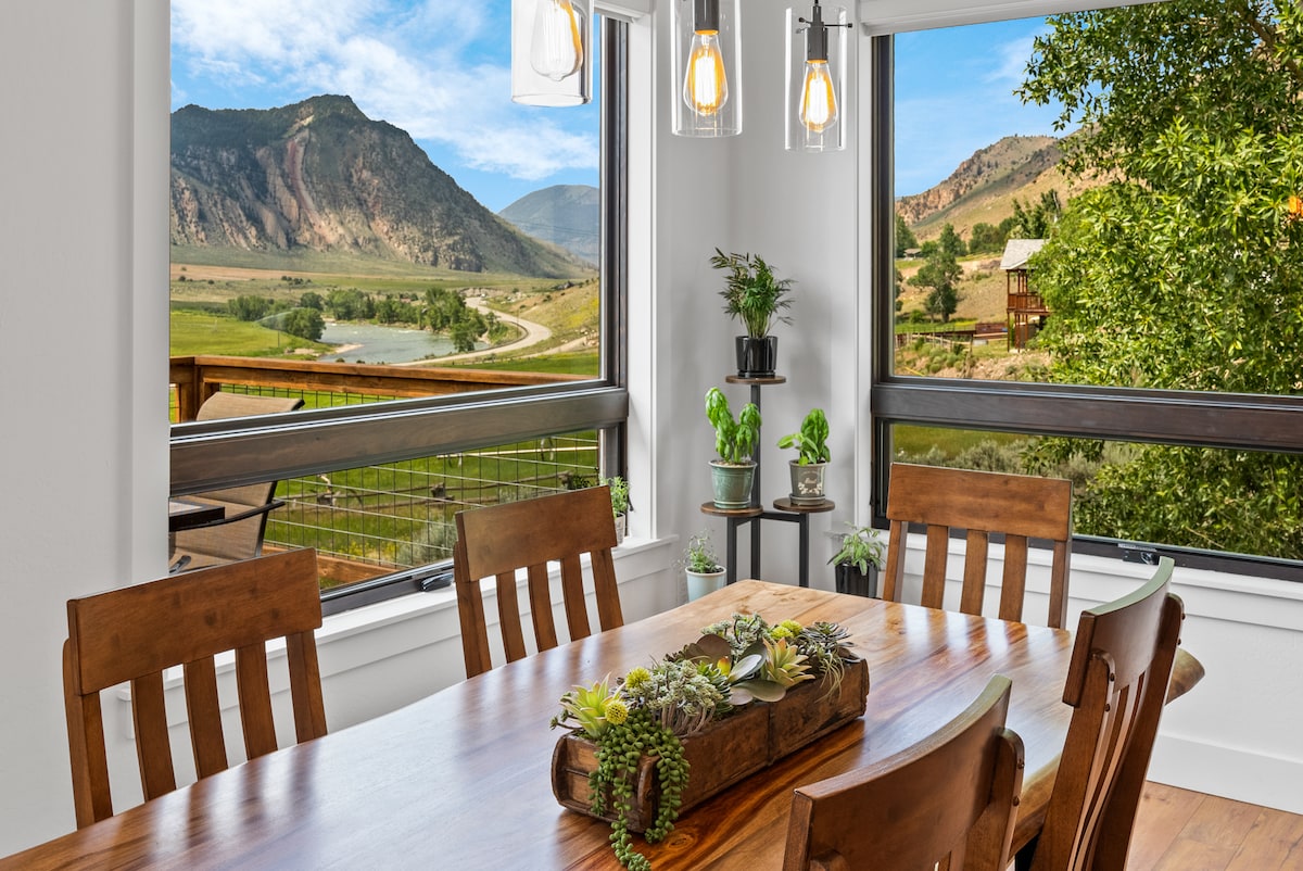 A dining area features a wooden table surrounded by sturdy chairs, set against expansive windows showcasing stunning mountain views. Natural light illuminates the space, while a greenery centerpiece adds a touch of nature to the setting.