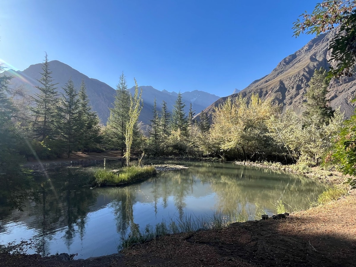 A serene mountain lagoon is surrounded by lush greenery and tall trees, reflecting the clear blue sky and surrounding mountains. The tranquil waters highlight the natural beauty of the area, with gentle ripples visible on the surface.