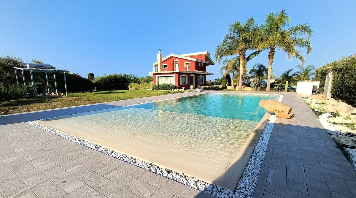 A large, inviting swimming pool is set within a spacious outdoor area, surrounded by palm trees and manicured gardens. The villa, characterized by bold red exterior, stands in the background, enhancing the tranquil ambiance of the property.