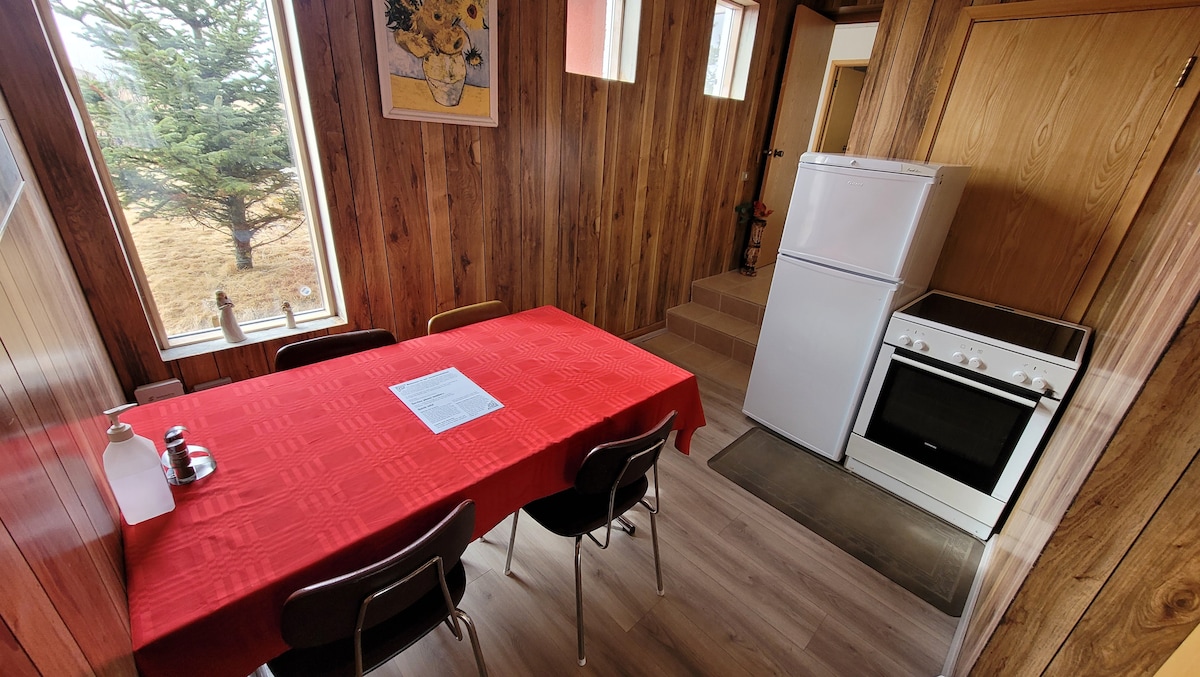 A dining area features a red tablecloth on a rectangular table surrounded by chairs. A white fridge and stove are positioned in the kitchen space, with wooden paneling creating a cozy atmosphere. Light enters through two windows, highlighting the simplicity of the room.