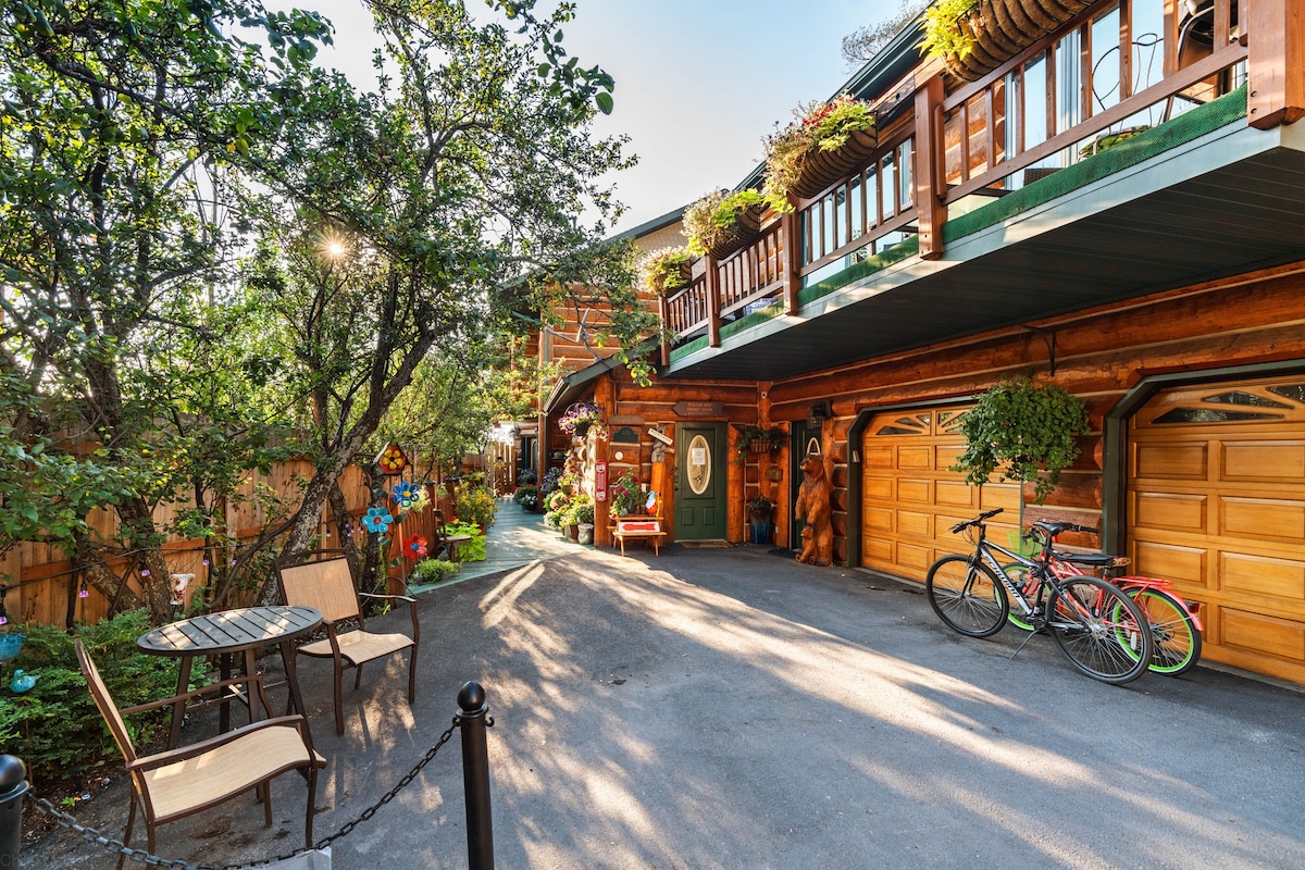 A charming outdoor area is presented, showcasing a paved driveway flanked by vibrant greenery and decorative plants. Two bicycles are parked nearby, and a small table with chairs is nestled under the shade of trees, inviting relaxation in the serene environment.