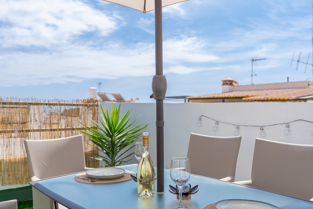 A private terrace is shown, featuring a table set for dining under a large umbrella. Two beige chairs surround the table, complemented by a bottle of wine and a glass. A potted plant adds a touch of greenery, while string lights are visible in the background.