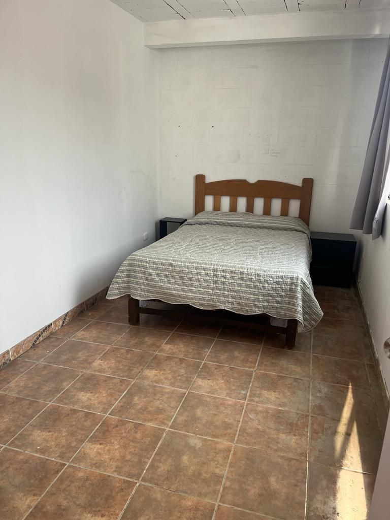 A simple sleeping area features a wooden bed with a patterned blanket placed atop. Neutral-colored walls provide a calm backdrop, while a gray curtain frames a window allowing natural light to enter. The tiled floor adds a touch of warmth to the space.
