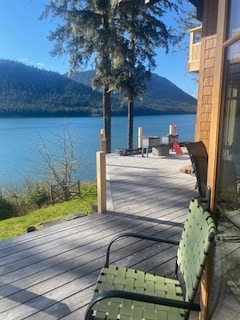 A wooden deck extends towards the water, providing a view of the lake framed by green trees and mountains in the background. A single green chair is positioned to enjoy the scenery, with additional seating and fire pit visible nearby.