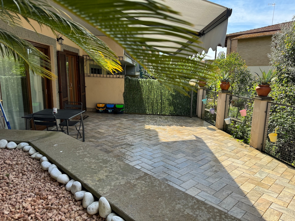 A spacious outdoor patio is shown, featuring a shaded area beneath a retractable awning. Potted plants line the perimeter, and colorful planters are visible on the fence. A black folding table and chairs are situated on the tiled surface, providing a place to relax.