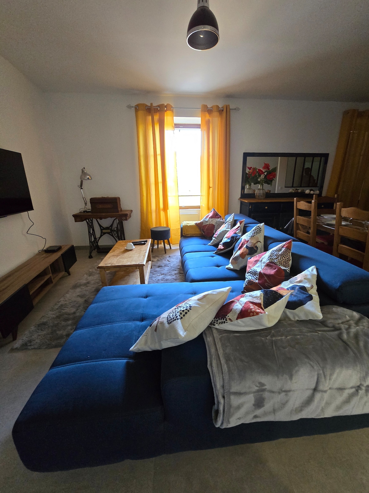A spacious living area features a large blue sectional sofa adorned with decorative pillows. A coffee table is placed in front, complementing a flat-screen television mounted on the wall. Sunlight filters through yellow curtains, illuminating the room and a nearby dining area with wooden chairs.