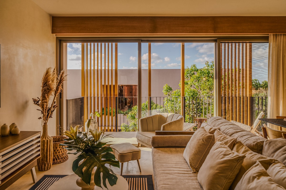 A bright and airy living area is presented, featuring large glass doors that open to a balcony. Natural light floods the space, accentuating the neutral tones of the furnishings, including a comfortable sofa and stylish decor. Lush greenery is visible outside, enhancing the tranquility of the setting.