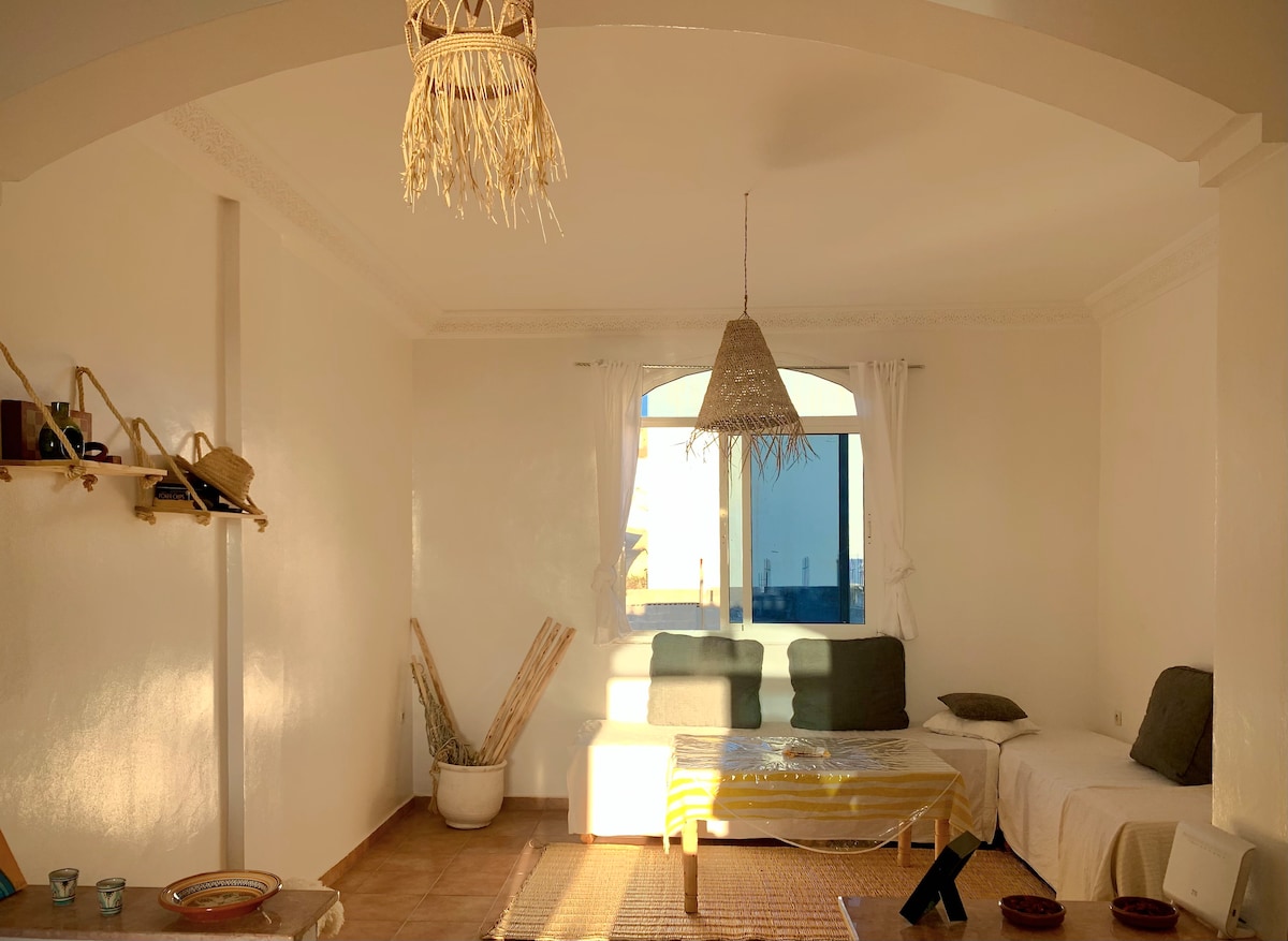 A bright living area features a large window allowing natural light to fill the space. Simple seating arrangements include a sofa and cushions, with a woven pendant light hanging above. Decorative elements such as a shelf with assorted items are visible against the white walls.