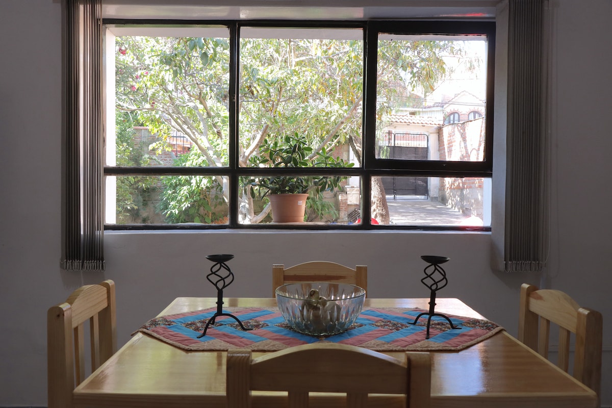 A light-filled dining area features a wooden table surrounded by four chairs. A colorful woven table runner rests in the center, adorned with a glass bowl. Large windows provide a view of greenery outside, enhancing the relaxed atmosphere of the space.
