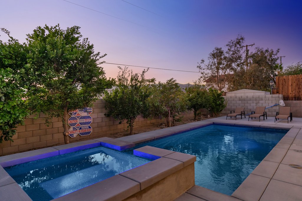 A serene outdoor space features a swimming pool and spa, set against a backdrop of soft desert landscaping. Lounge chairs sit nearby, and illuminated blue water creates a calming atmosphere as twilight approaches.