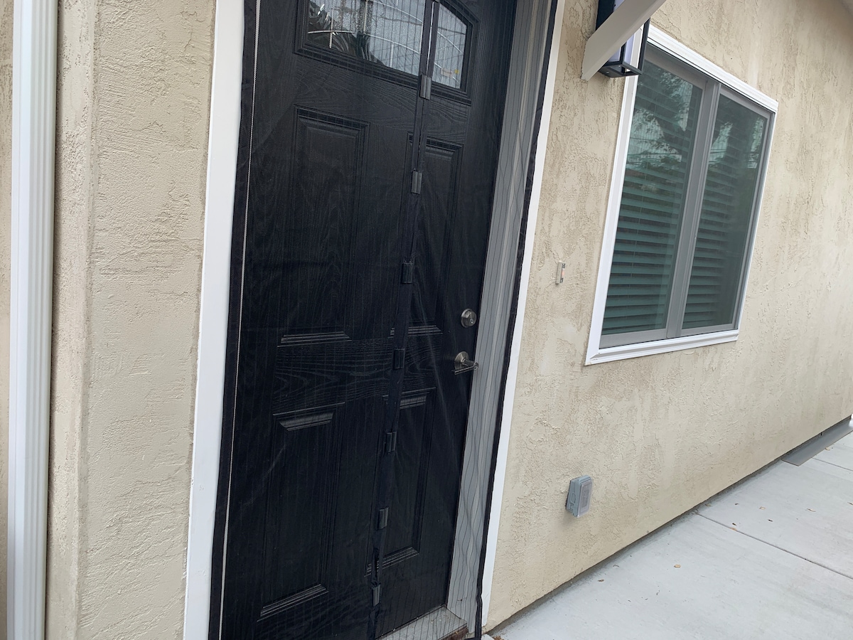 The image displays the entrance of the studio, featuring a solid black door with vertical paneling. Natural light is visible through adjacent windows with divided sections. A smooth concrete pathway leads to the entrance, framed by a textured beige wall.