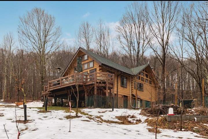 The cabin features a contemporary log design, with large windows and a spacious wrap-around deck. Surrounded by trees, the structure stands on a snowy landscape, showcasing a connection to nature. A landscaped area is visible, enhancing the serene setting.