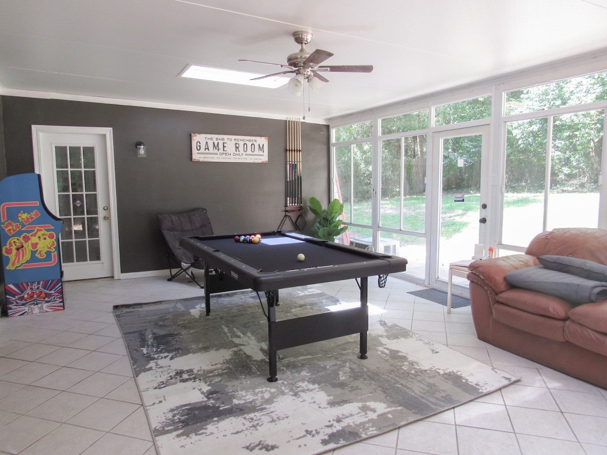 A spacious game room features a 7ft pool table, two couches, and arcade games. Large windows allow natural light to fill the space, highlighting the natural surroundings visible through the glass doors. The room is decorated with a neutral area rug and includes a ceiling fan.