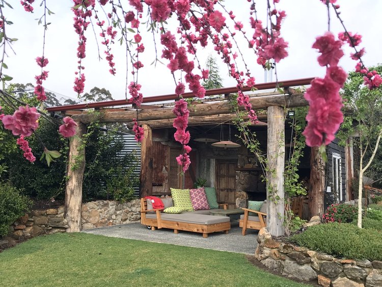 An outdoor seating area is framed by flowering branches, providing a vibrant contrast against natural wooden beams. Comfortable lounge furniture with decorative cushions is arranged on a stone patio, complemented by a landscaped garden in the background.