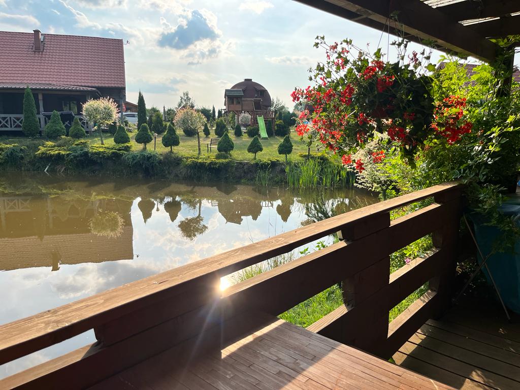 A serene view of a well-kept pond reflects the surrounding greenery and nearby structures. Vibrant red flowers hang from the wooden deck, enhancing the natural setting. The calm water captures the essence of the tranquil atmosphere, inviting relaxation in the surrounding landscape.