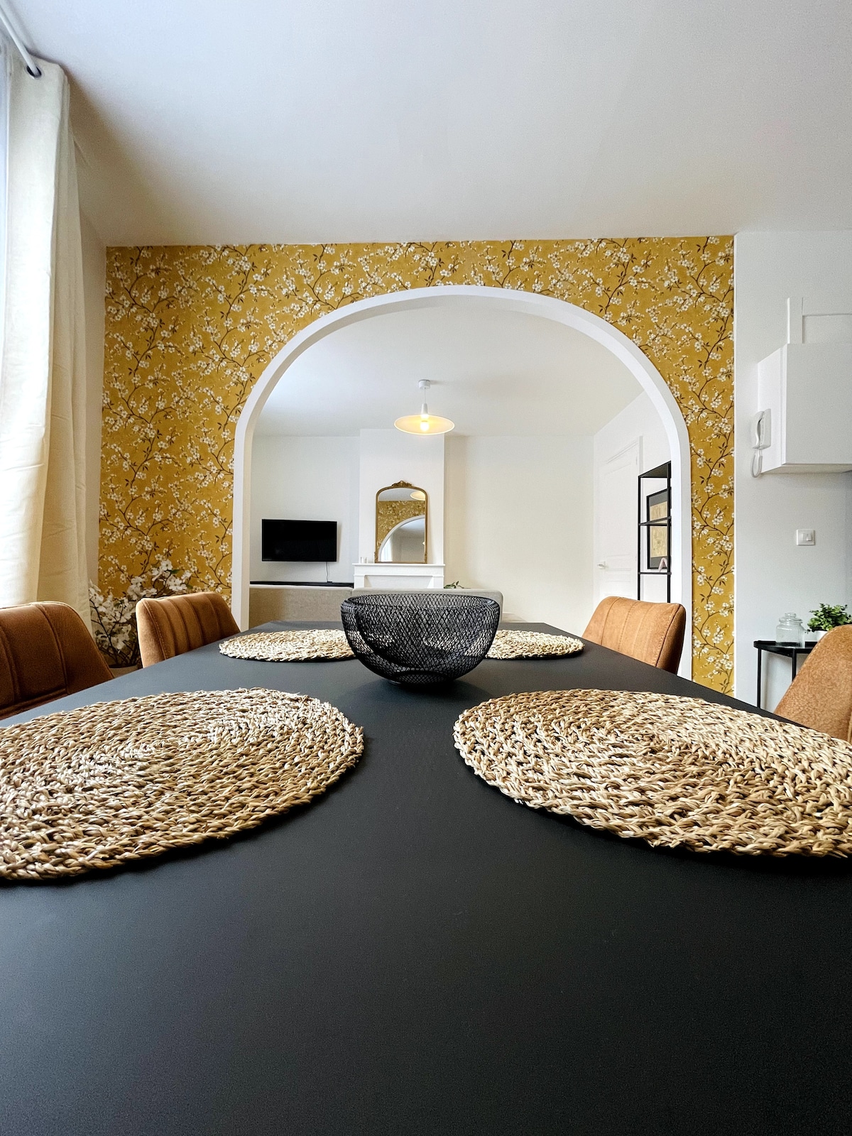 A dining area is presented with a long black table, surrounded by brown chairs. Natural fiber placemats are neatly arranged on the table. An archway leads to a bright living space, which features a decorative mirror and a television in the background, alongside a light-coloured wall.