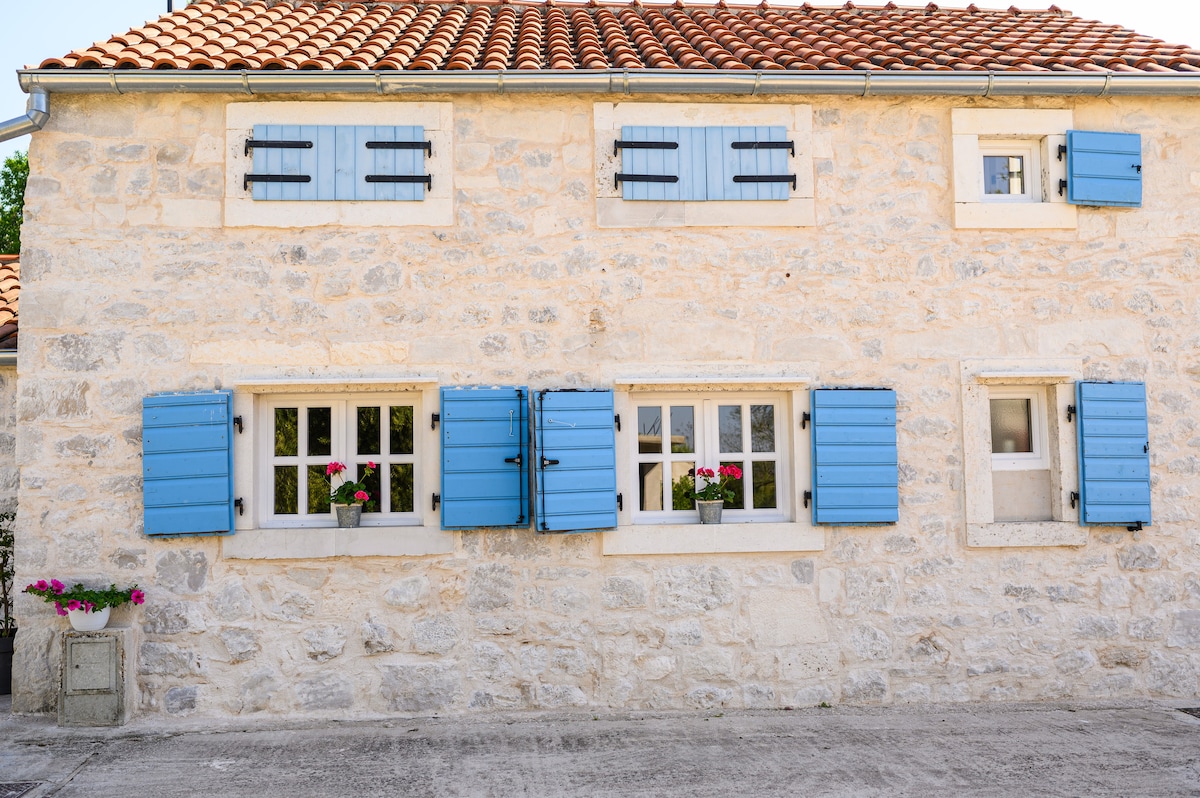 The exterior features a stone facade accompanied by bright blue shutters that enhance the charm of the house. Flower boxes are positioned beneath the windows, adding natural color to the design. A traditional tile roof complements the overall aesthetic.