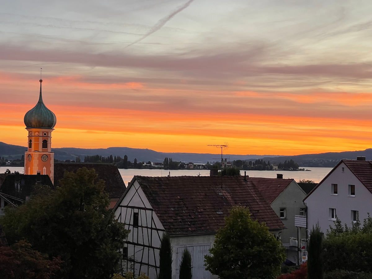 A vibrant sunset casts shades of orange and pink across the sky, reflecting on the calm waters. In the foreground, rooftops of charming houses can be seen, while a distinct tower with a green dome stands prominently against the backdrop of the scenic landscape.