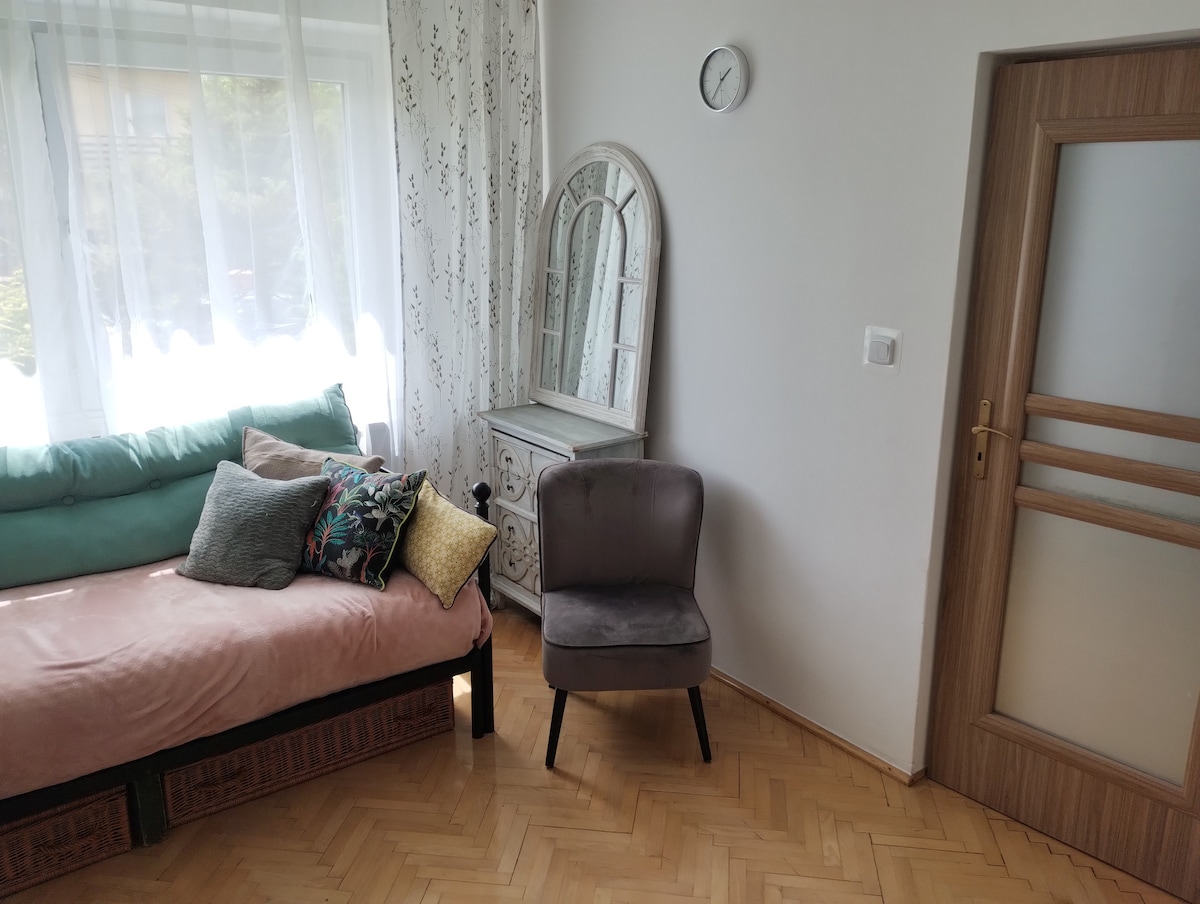A cozy sitting area features a daybed adorned with decorative pillows alongside a plush chair. A vintage mirror is positioned above a small dresser, with natural light filtering through sheer curtains. A simple wall clock adds a touch of functionality to the space.