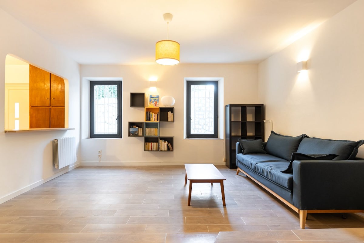 A bright and airy living area features a comfortable dark sofa and a wooden coffee table set on light tile flooring. Two windows with grid-patterned panes allow natural light to fill the space. A shelf displays books against a neutral backdrop, enhancing the room's simplicity.