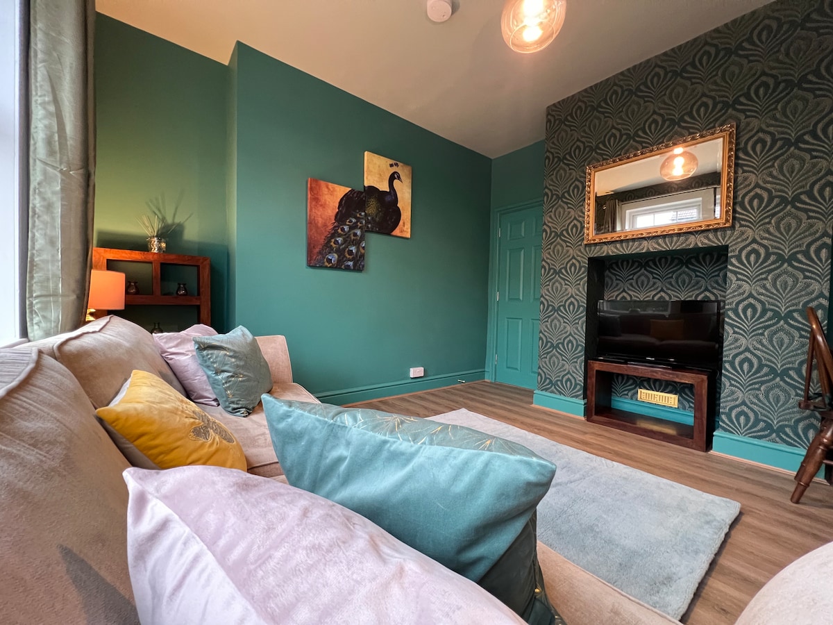 A cozy living area features a chenille sofa adorned with decorative cushions in various shades. A soft area rug rests on the floor, while a wall display includes a peacock artwork. A flat-screen TV is positioned on a dark wood console against an elegantly patterned wall.