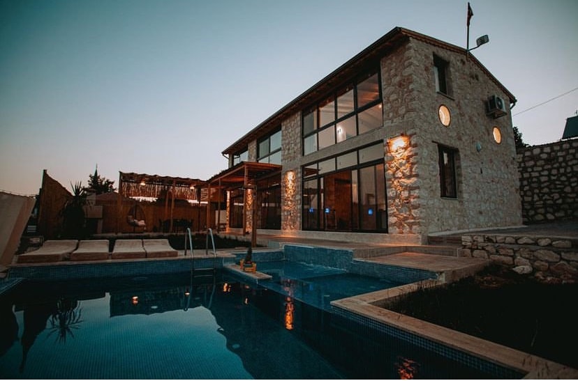 A spacious stone house is highlighted by large windows that reflect the evening light. A serene swimming pool is positioned in the foreground, with wooden deck chairs arranged nearby. The outdoor area features a shaded seating space, inviting relaxation.