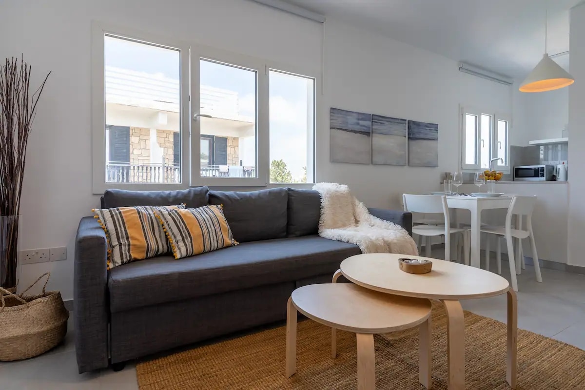 A modern living area features a grey sofa adorned with striped cushions, complemented by a cozy white throw. Two light wooden coffee tables sit on a woven area rug. Natural light enters through three large windows, illuminating a dining table and kitchenette in the background.